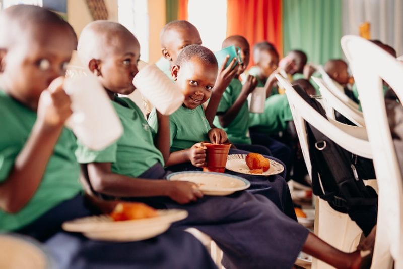 alt="Ein Tag im Compassion Kinderzentrum in Ruanda. Kinder sitzen mit Essen auf Plastikstühlen und essen Brötchen und Trinken Tee."