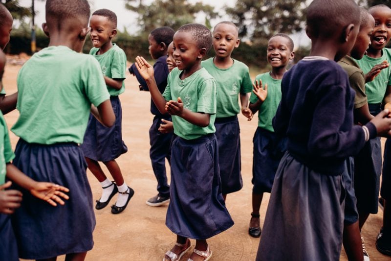 alt="Ein Tag im Compassion Kinderzentrum in Ruanda. Kinder tanzen auf dem Hof des Kinderzentrums und freuen sich dabei. Sie tragen Schuluniform."