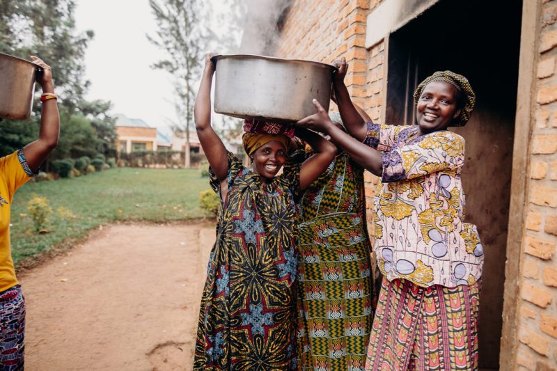 alt="Ein Tag im Compassion Kinderzentrum in Ruanda. Mütter, Kirchenmitarbeiter und freiwillige Helfer stehen zusammen und tragen eine große Schüssel mit warmen Essen"