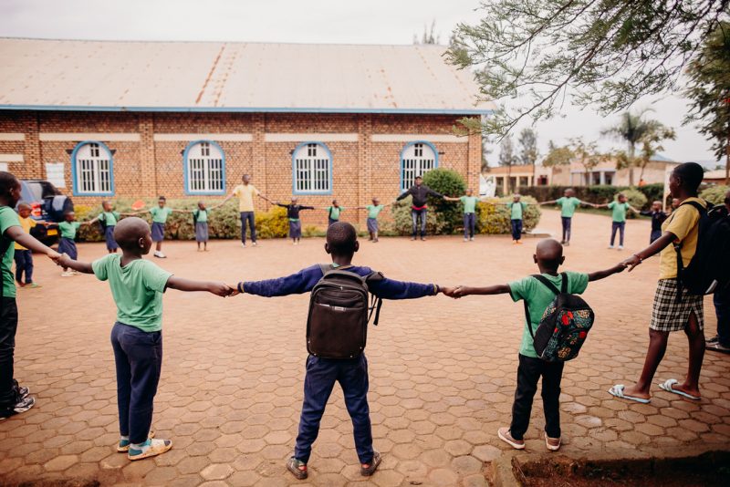 alt="Ein Tag im Compassion Kinderzentrum in Ruanda. Kinder stehen im Kinderzentrum zusammen und bilden einen großen Kreis, Hand in Hand."