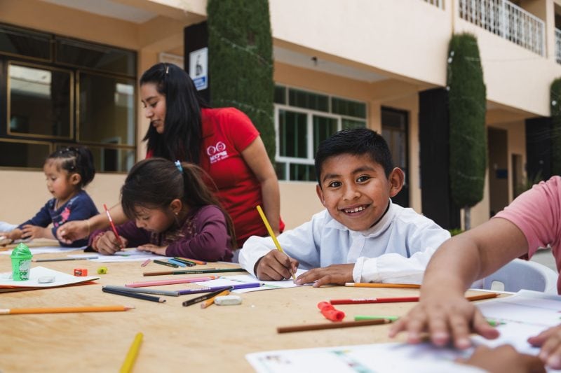 alt="Warum stehen Kinder in Armut bei Compassion im Blickpunkt? Kinder im Blickpunkt, Kinder sitzen an einem Schreibtisch in einem Kinderzentrum und malen mit Buntstiften"