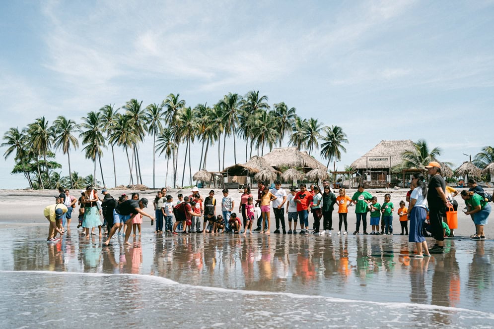 alt="Kinder schützen Meeresschildkröten, Ruben, El Salvador, Kinder stehen in einer Reihe am Strand und lassen Schildkröten frei, Palmen wedeln im Wind und Häuser stehen am Strand, Compassion Deutschland"