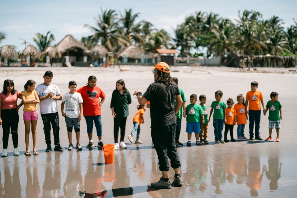 alt="Kinder schützen Meeresschildkröten, Ruben, El Salvador, Ruben lässt am Strand Schildkröten frei, Kinder stehen an einer Reihe, Palmen wedeln im Wind und Häuser stehen am Strand, Compassion Deutschland"